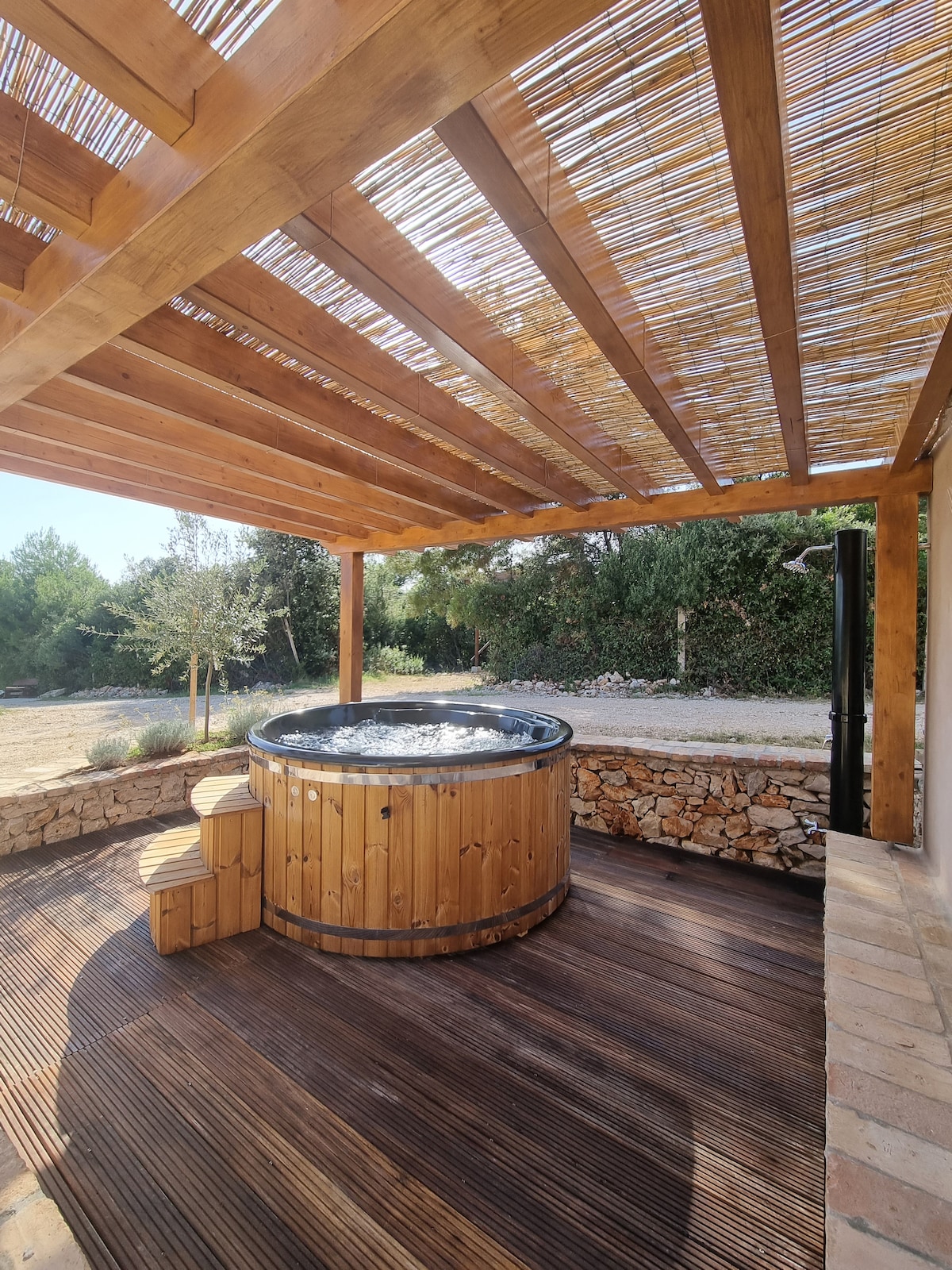 A wooden hot tub is situated under a slatted pergola, surrounded by natural greenery. The deck features circular wooden seating for comfort. Soft sunlight filters through the overhead structure, highlighting the smooth textures of the tub and the warm tones of the wooden flooring.