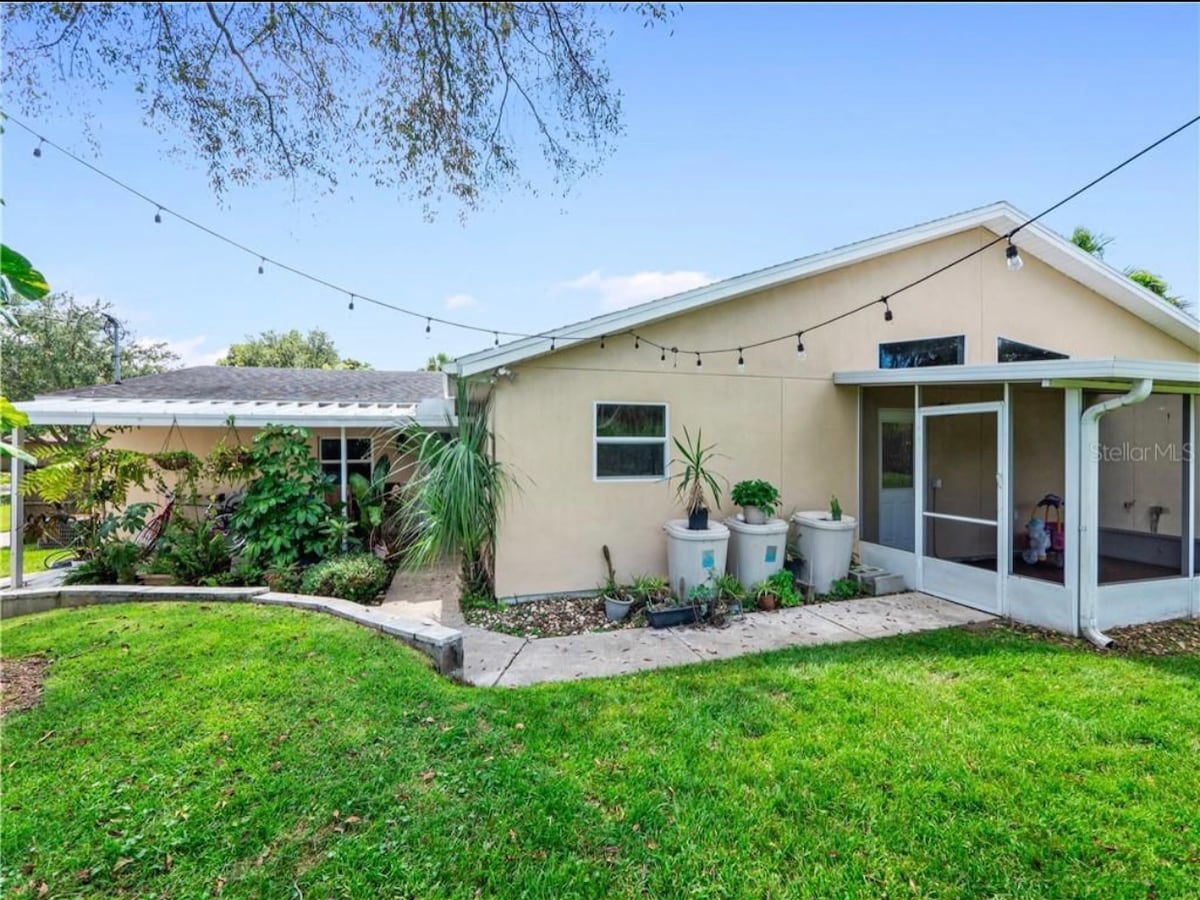 The exterior of the home features a patio area surrounded by lush greenery and potted plants. String lights are hung above, creating an inviting atmosphere. The pathway is lined with manicured grass leading to a covered entryway.
