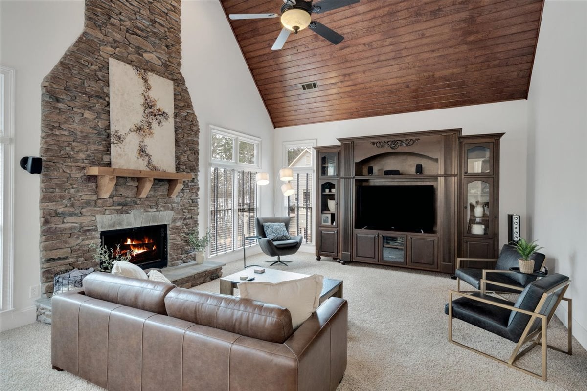 A spacious living room features a stone fireplace beside large windows, allowing natural light to fill the area. A comfortable leather sofa faces a dark wood entertainment center, while a modern chair and potted plant add inviting touches to the decor.