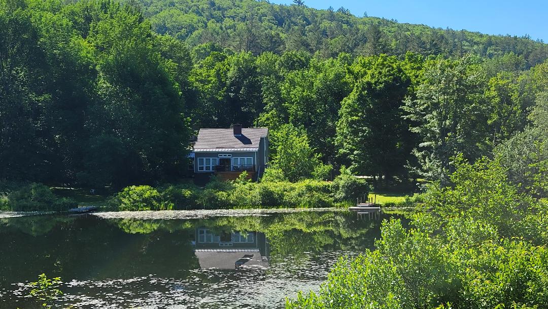 A serene view of the lakefront house, surrounded by lush greenery and trees. The reflective water showcases a calm atmosphere, with the house's structure and soft colors blending harmoniously with the natural landscape.