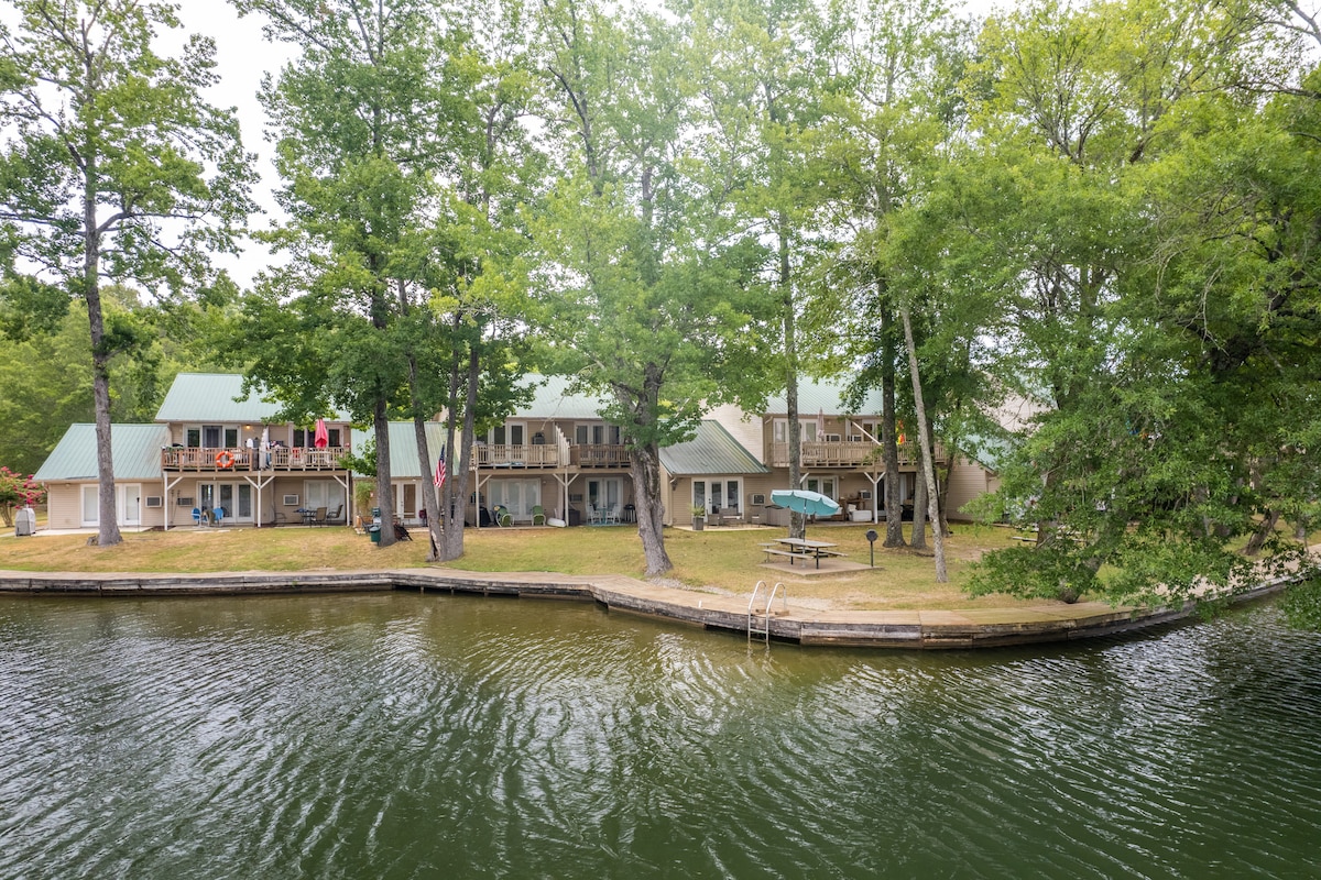 The exterior of the condo complex is surrounded by lush trees, providing a shaded and tranquil setting by the water. A grassy area lies adjacent to the lake, with a private dock extending into the calm water, which reflects the greenery and buildings.