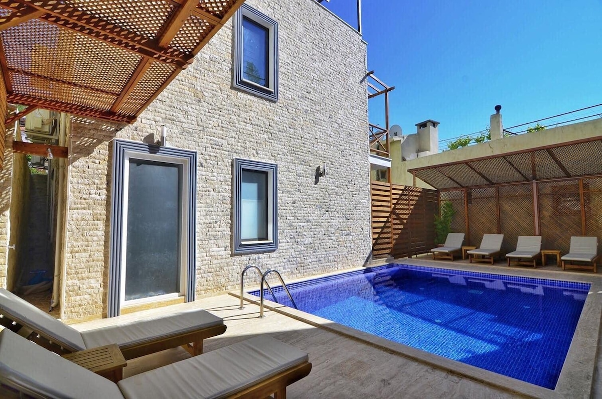 A private pool area is depicted, featuring a tiled pool surrounded by a stone deck. Six loungers are arranged for relaxation. The villa's exterior showcases a mix of textured stone and large windows, while a shaded seating structure offers additional comfort.