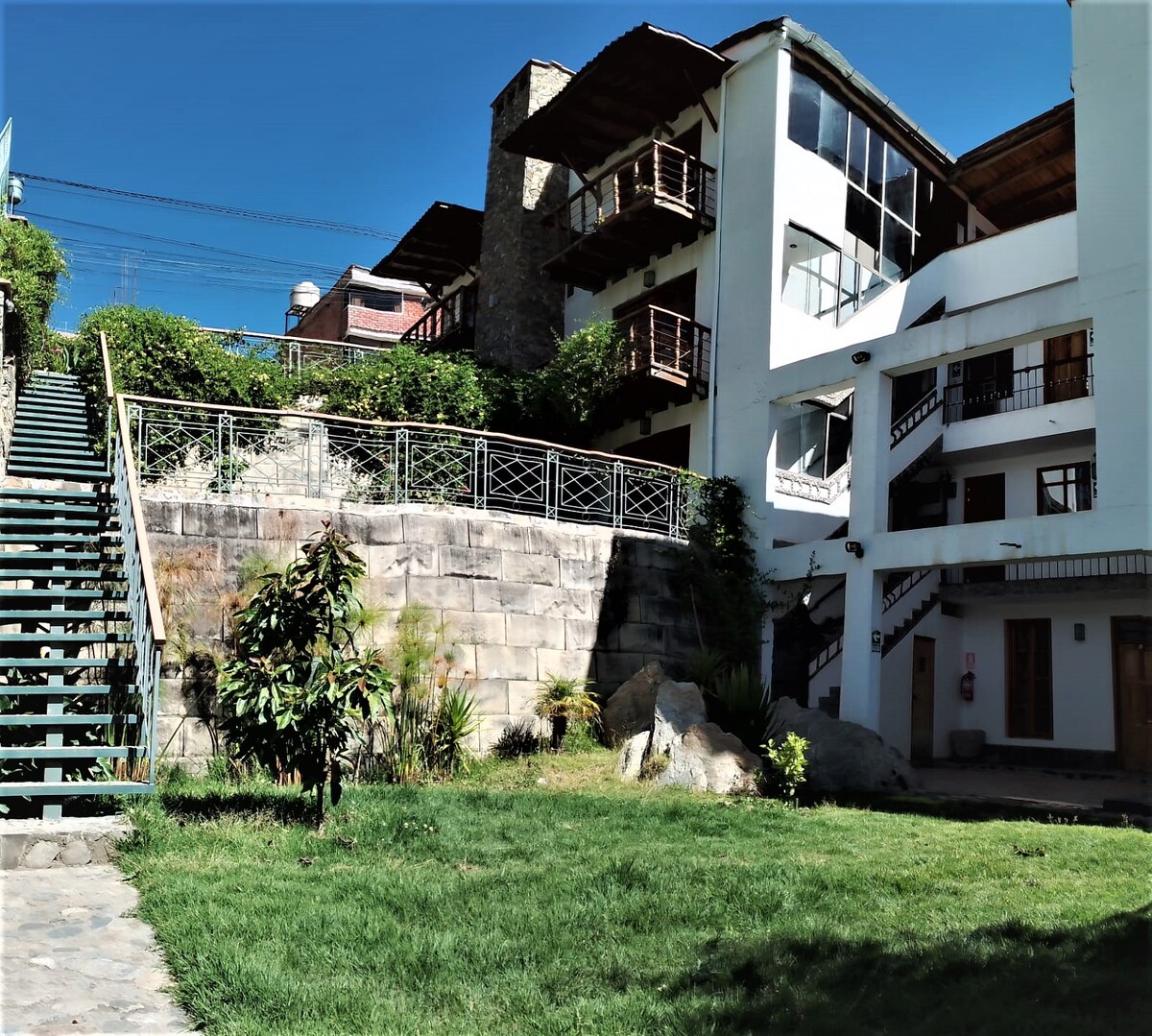 A well-maintained garden area features lush green grass and plants, bordered by natural stone. The multi-story building is visible in the background, with balconies on upper levels. A set of stairs leads from the garden to the entrance of the structure, providing access to the rooms.