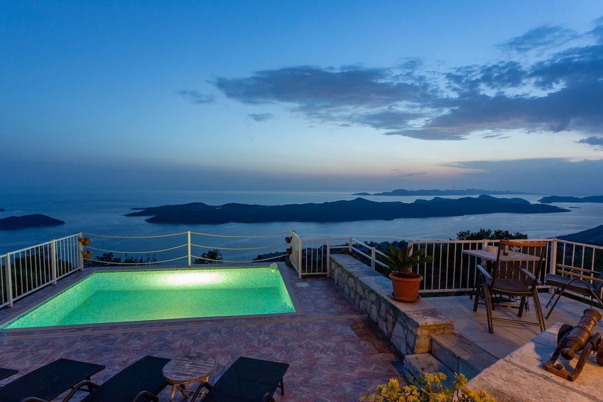 A glistening swimming pool is positioned on a terrace, offering views of distant islands and the horizon at twilight. Surrounding areas are adorned with lounge chairs and greenery, creating a tranquil environment perfect for relaxation.