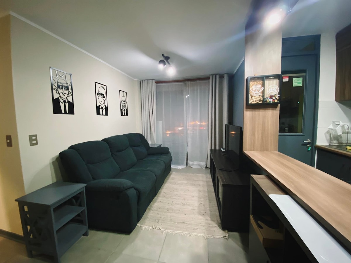 A spacious living area features a dark green sofa positioned near a large window, allowing for natural light to illuminate the room. Two framed artworks adorn the walls, and a light-colored area rug complements the tile flooring.