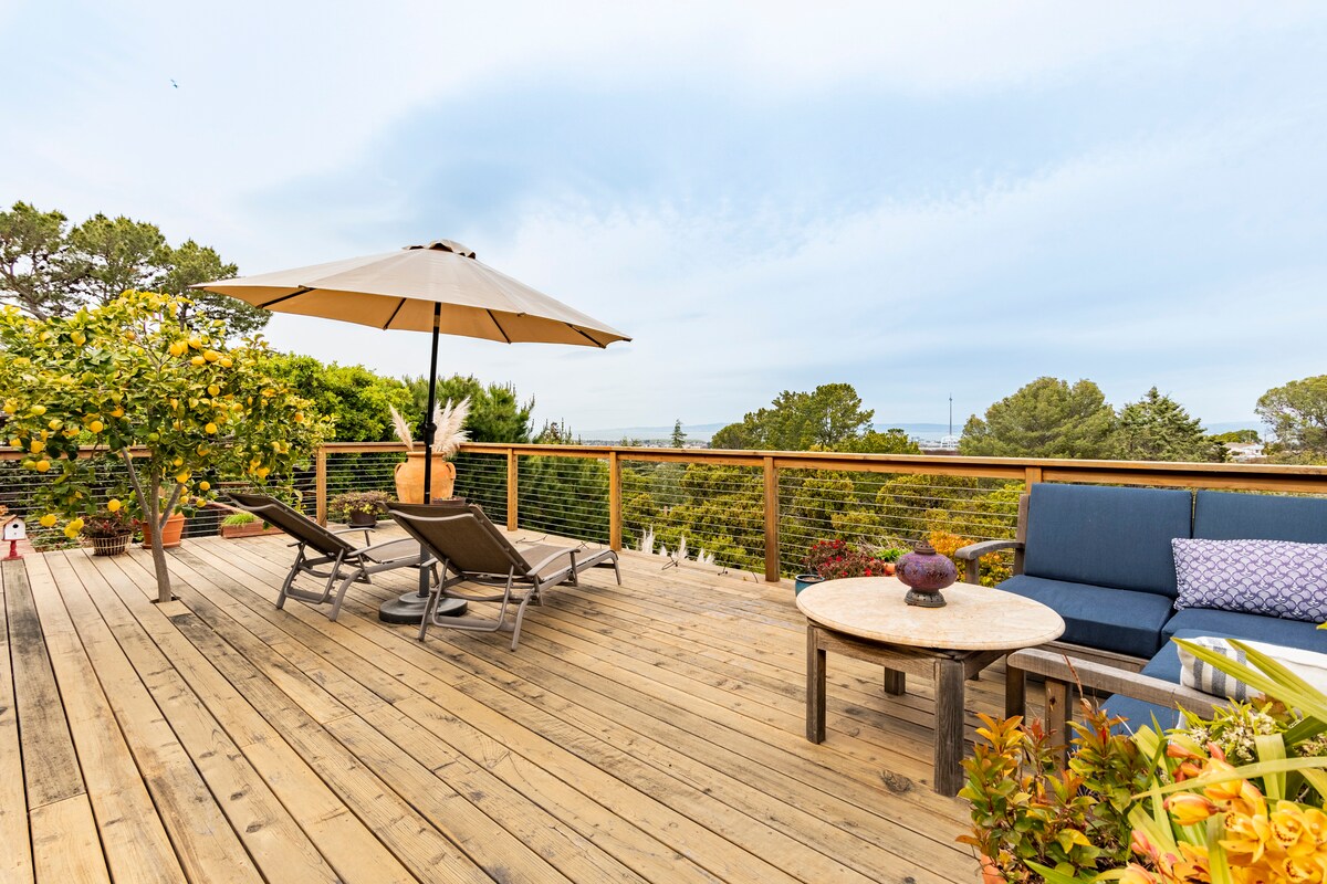 A spacious outdoor patio features wooden decking and a large umbrella providing shade. Comfortable lounge chairs are complemented by a sectional couch with plush cushions. Vibrant potted plants and a view of greenery and distant hills enhance the serene atmosphere.