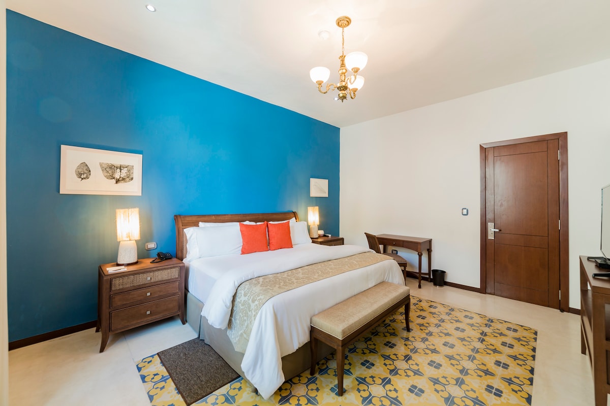 A spacious bedroom features a large bed with soft linens and decorative pillows. A bright blue accent wall contrasts with neutral tones, while a patterned area rug adds texture. Two bedside lamps illuminate the space, which includes a wooden desk and chair in the corner.