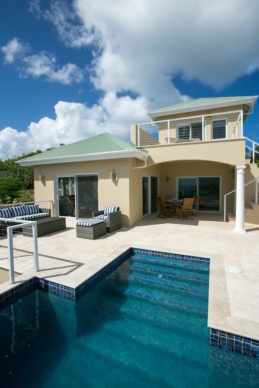 The exterior view of the cottage showcases a private plunge pool with stone steps leading in. Outside seating is provided on a coral stone deck, adjacent to large glass doors that open into the living area. A second story is visible, offering additional outdoor space.