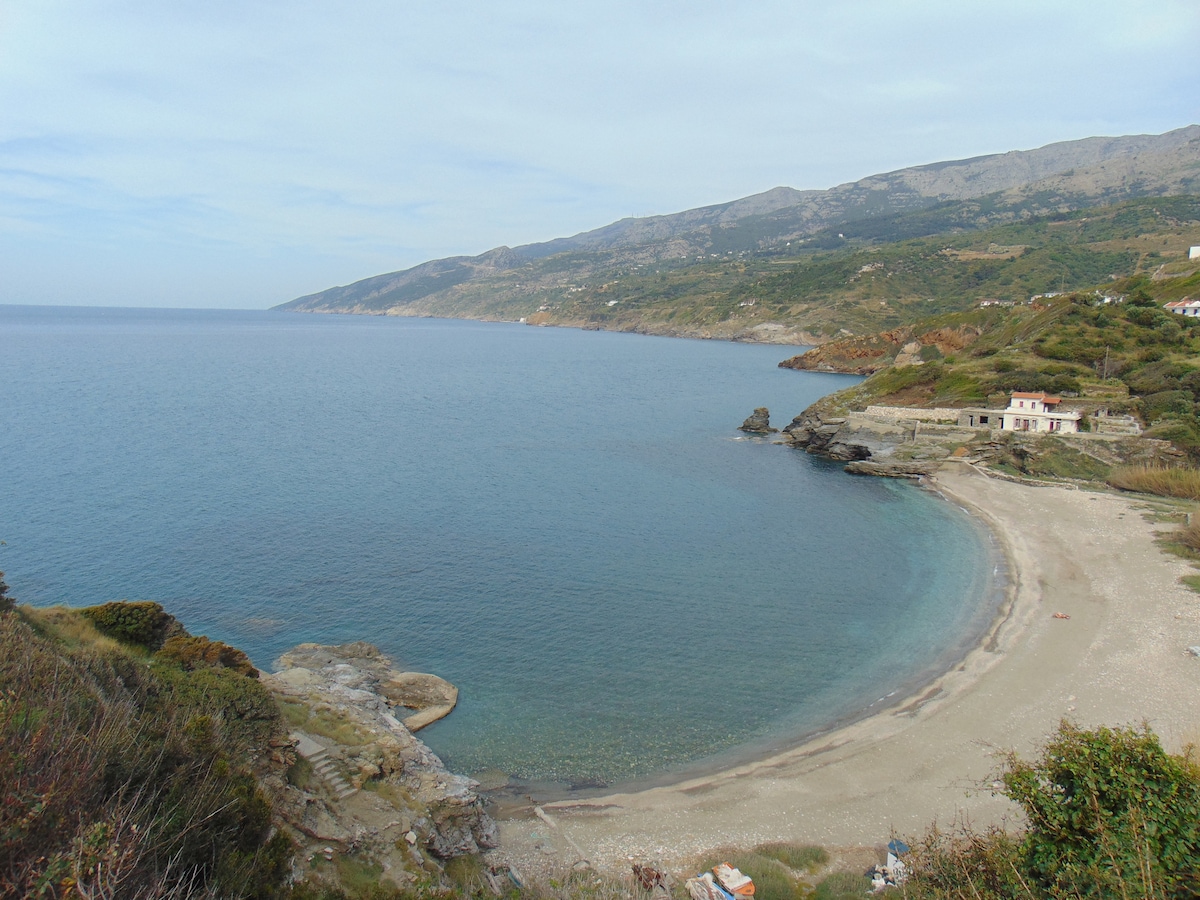 A serene bay is depicted, featuring a gentle curve of sandy shoreline bordered by clear blue waters. Rolling hills and a mountain range are visible in the background, adding to the tranquil landscape. A few buildings are nestled along the shore, blending with the natural surroundings.