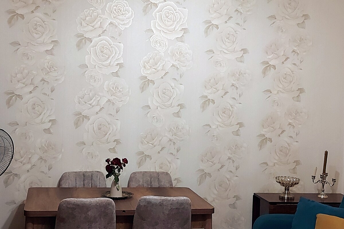 A dining area is presented with a wooden table surrounded by four upholstered chairs. The wall features a delicate floral design, with large white roses creating a serene backdrop. A small vase with flowers is placed at the center of the table, adding a subtle touch of elegance.