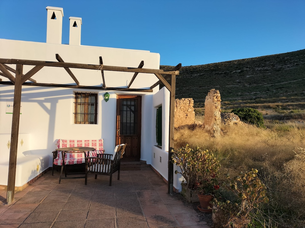 A charming outdoor patio area features a wooden table with matching chairs, set against a backdrop of natural grasslands and distant hills. Sunlight illuminates the white exterior of the house, while a stone structure stands nearby, adding rustic character to the scene.