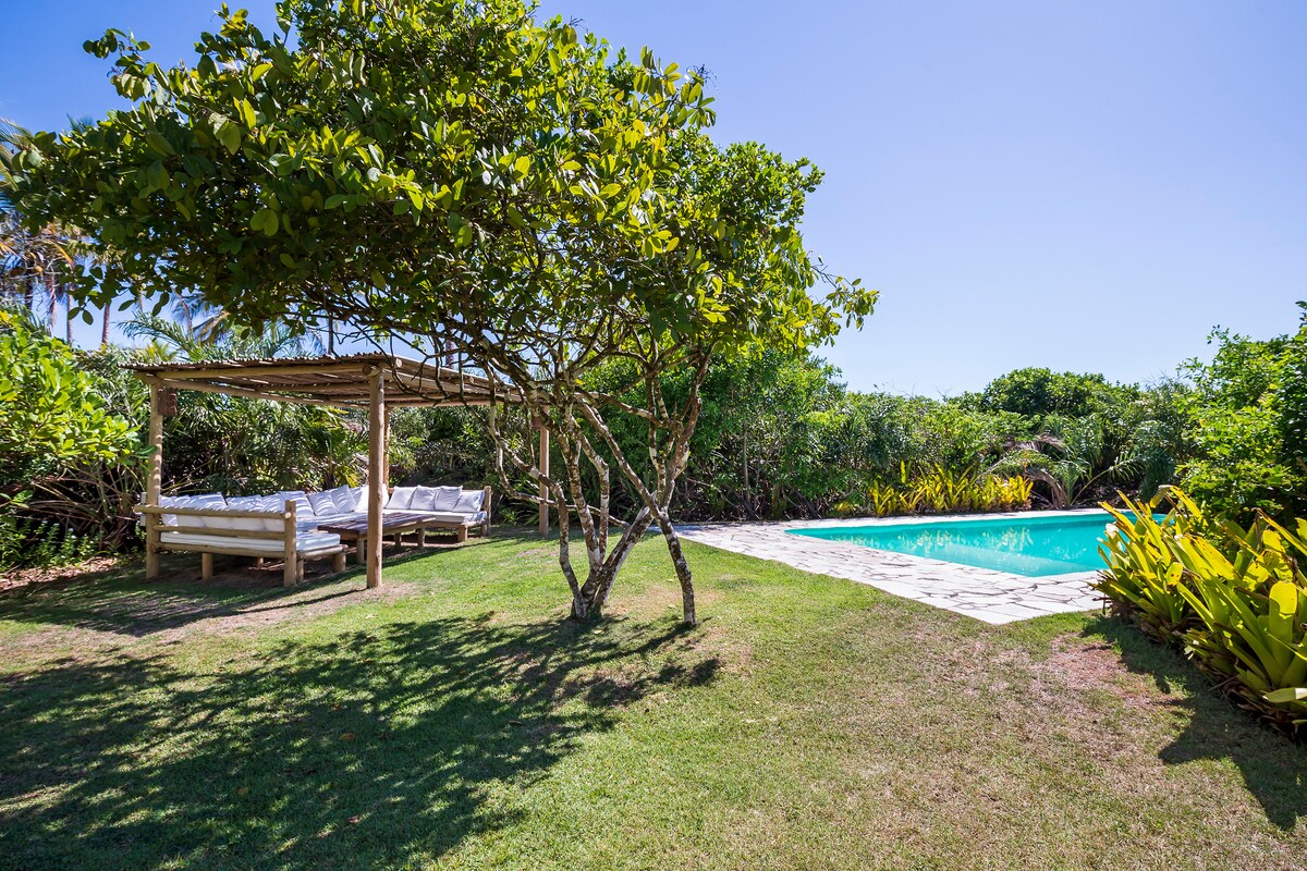 A shaded area is provided by a leafy tree, featuring a rustic gazebo with comfortable seating. Nearby, a swimming pool is seen, surrounded by lush green vegetation, offering a tranquil outdoor space for relaxation.