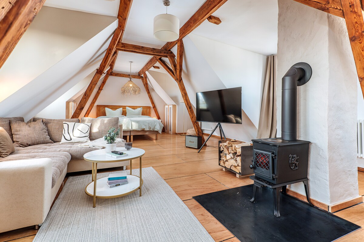 An open attic space features wooden beams and warm-toned decor. A spacious bed is positioned in the background, complemented by a flat-screen television mounted on the wall. A cozy sitting area includes a sofa, round coffee table, and a wood-burning stove, providing a homely ambiance.