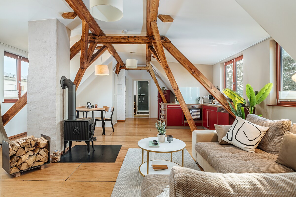 An airy living space is characterized by high, wooden beam ceilings and large windows allowing natural light. A cozy sofa is positioned near a modern coffee table, with a wood-burning stove nearby. The kitchen area features red cabinetry, and a dining table is visible in the background.