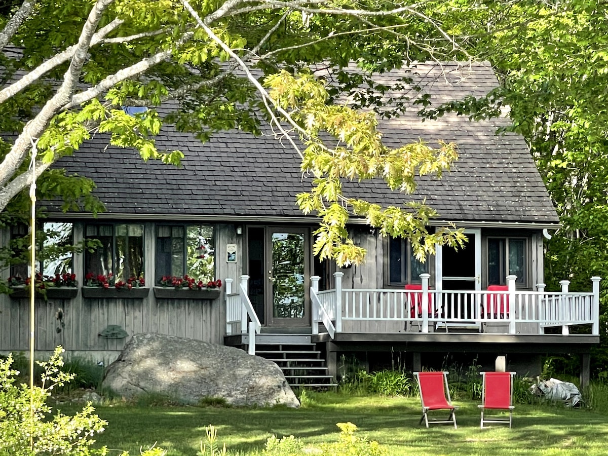 A charming cottage is nestled among greenery, featuring a welcoming porch with white railings and red chairs. Flower boxes adorn the windows, adding splashes of color. A large boulder and lush lawn provide a natural setting, enhancing the inviting atmosphere of the home's exterior.