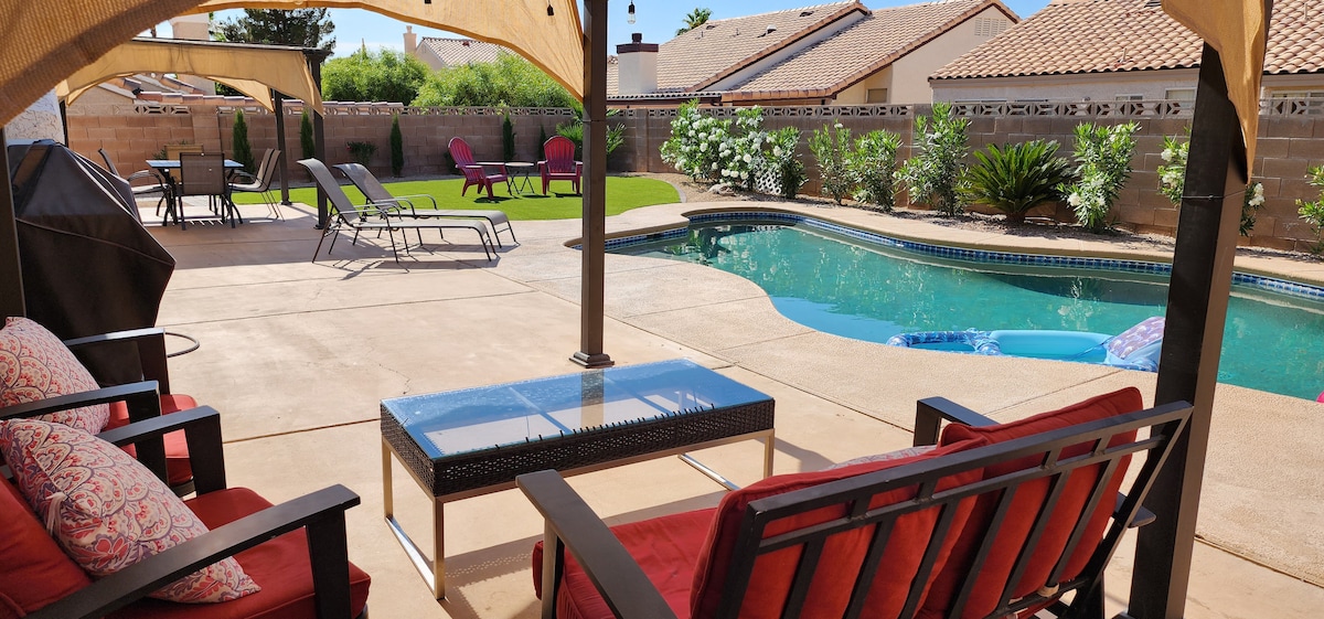 A private backyard features a sparkling pool and a spacious patio area. Comfortable seating is provided under a shaded canopy, with colorful cushions enhancing the inviting space. Lounge chairs are arranged around the pool, surrounded by lush greenery and soft landscaping.
