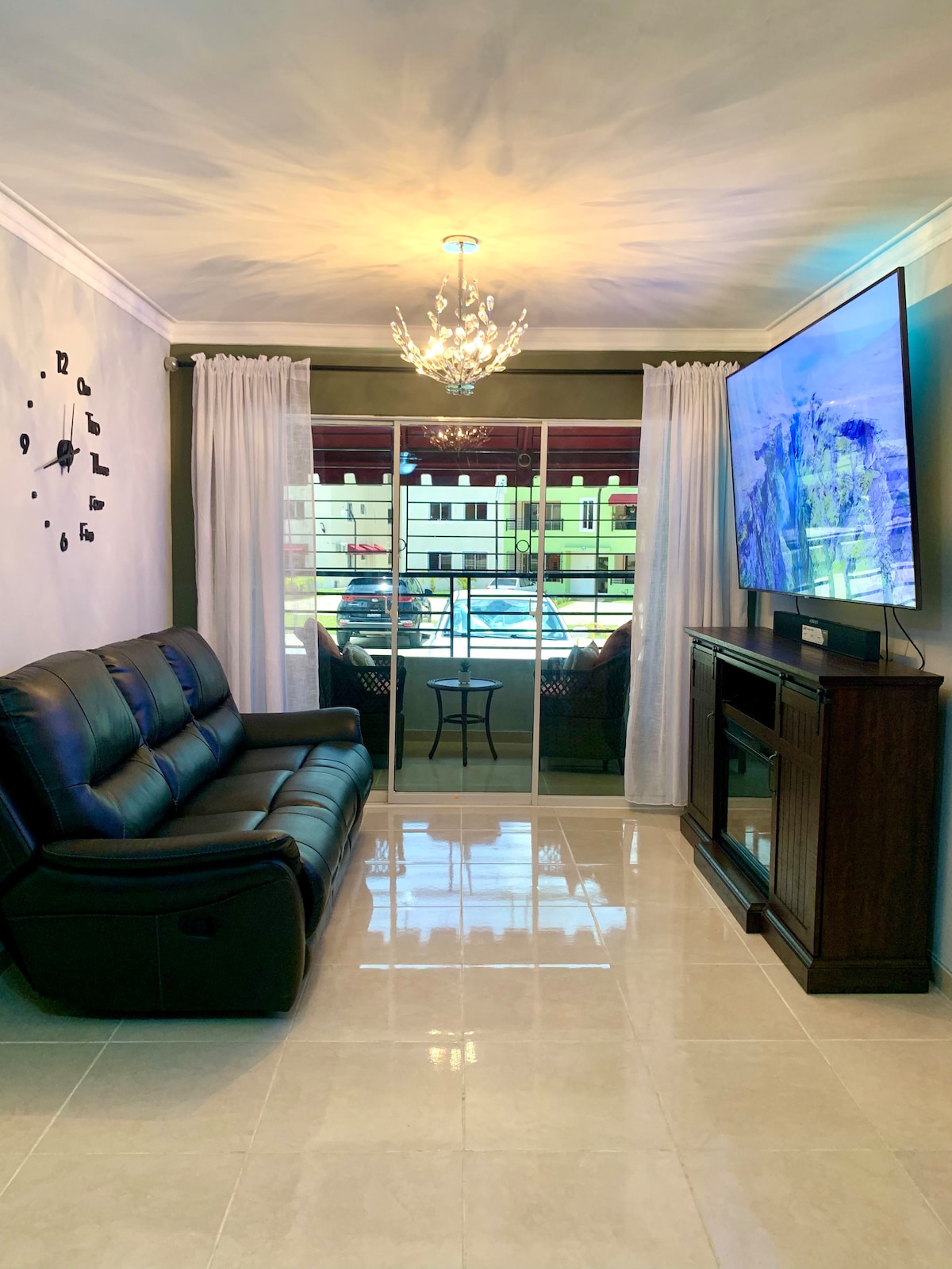 A living area showcases a sleek black leather sofa, complemented by a modern chandelier above. Floor-to-ceiling curtains frame a sliding glass door, leading to a balcony with an outdoor seating arrangement. A large flat-screen television is mounted on a stylish wooden console.