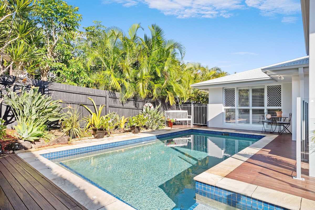 A private pool is framed by lush tropical plantings and a spacious wooden deck. Sunlight reflects off the water's surface, with a comfortable seating area visible to one side. The serene atmosphere is complemented by the modern home's expansive windows and light exterior.