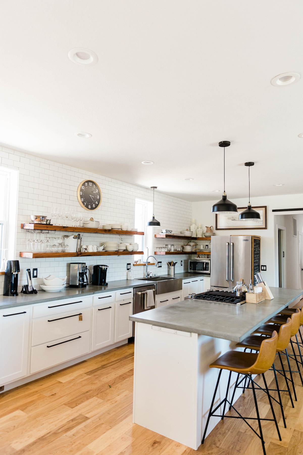 A modern kitchen features a large island with seating for several guests. White cabinetry and open shelving add functionality, with a clock displayed on the wall. Stainless steel appliances and a gas stove provide a contemporary touch, complemented by warm wooden flooring.