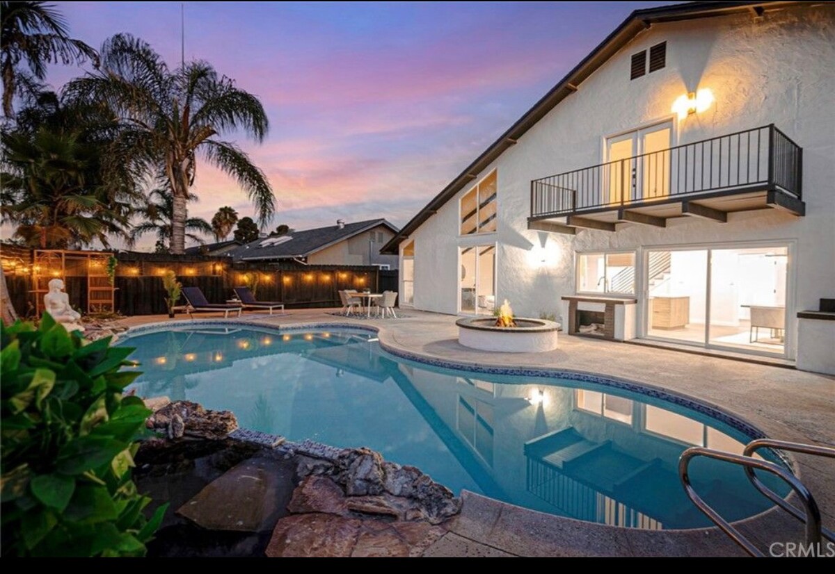 An inviting pool area is surrounded by palm trees and patio seating. The structure features large glass doors that provide a view of the interior. A fire pit is positioned beside the pool, all set against a colorful sunset sky.