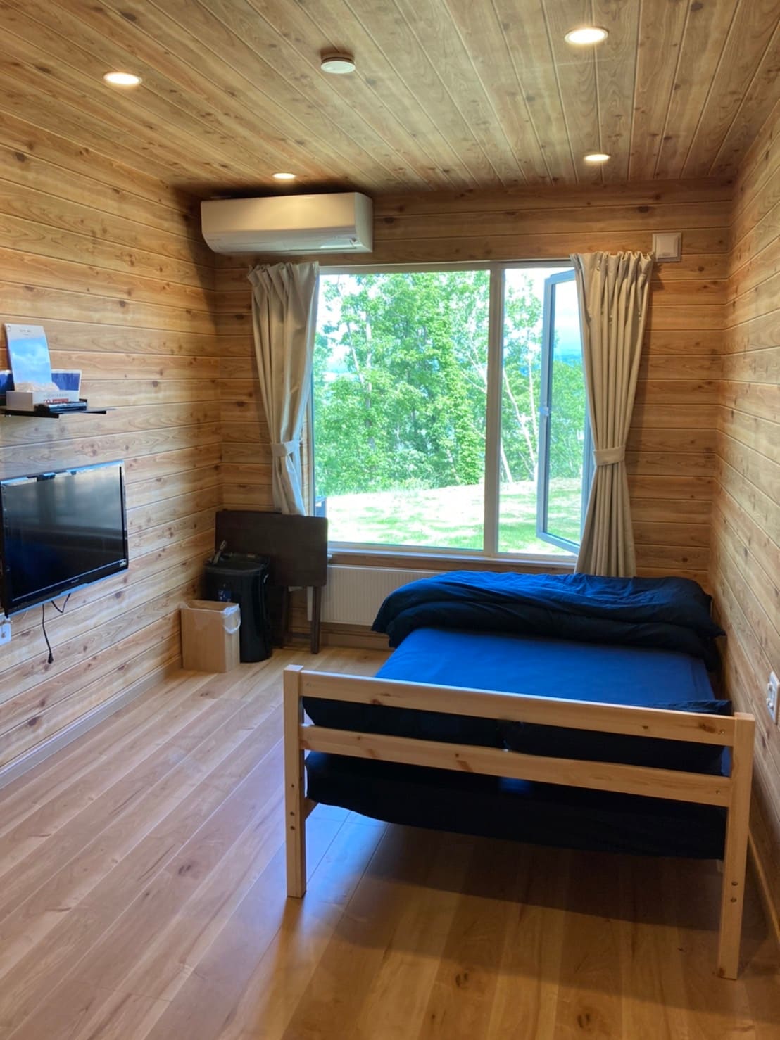 A room features wood-paneled walls and a large window overlooking greenery. A semi-double bed with blue bedding is complemented by a small desk and a television mounted on the wall. Natural light fills the space through the window, creating a bright environment.
