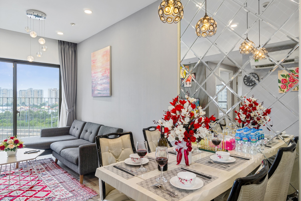 The dining area features a table set for six with elegant dinnerware and wine glasses. A stylish sofa is positioned nearby, complementing a colorful floral arrangement and artwork. Large windows offer a view of the city skyline, filling the space with natural light.