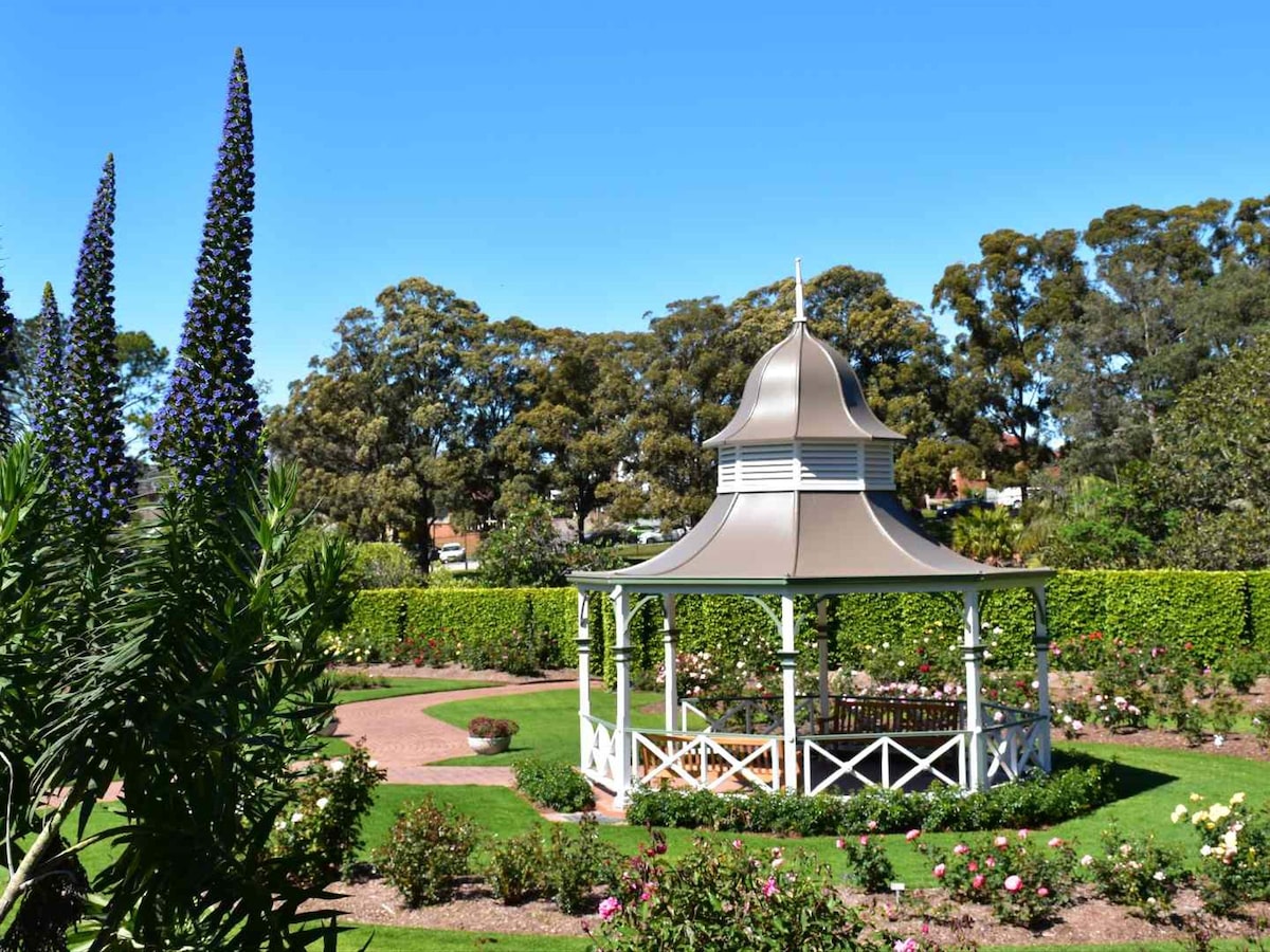 A traditional white gazebo is centered in a beautifully maintained garden, surrounded by colorful flower beds and lush greenery. Tall purple flowers add a pop of color in the foreground, while a well-trimmed hedge outlines the garden, creating a serene atmosphere for relaxation.