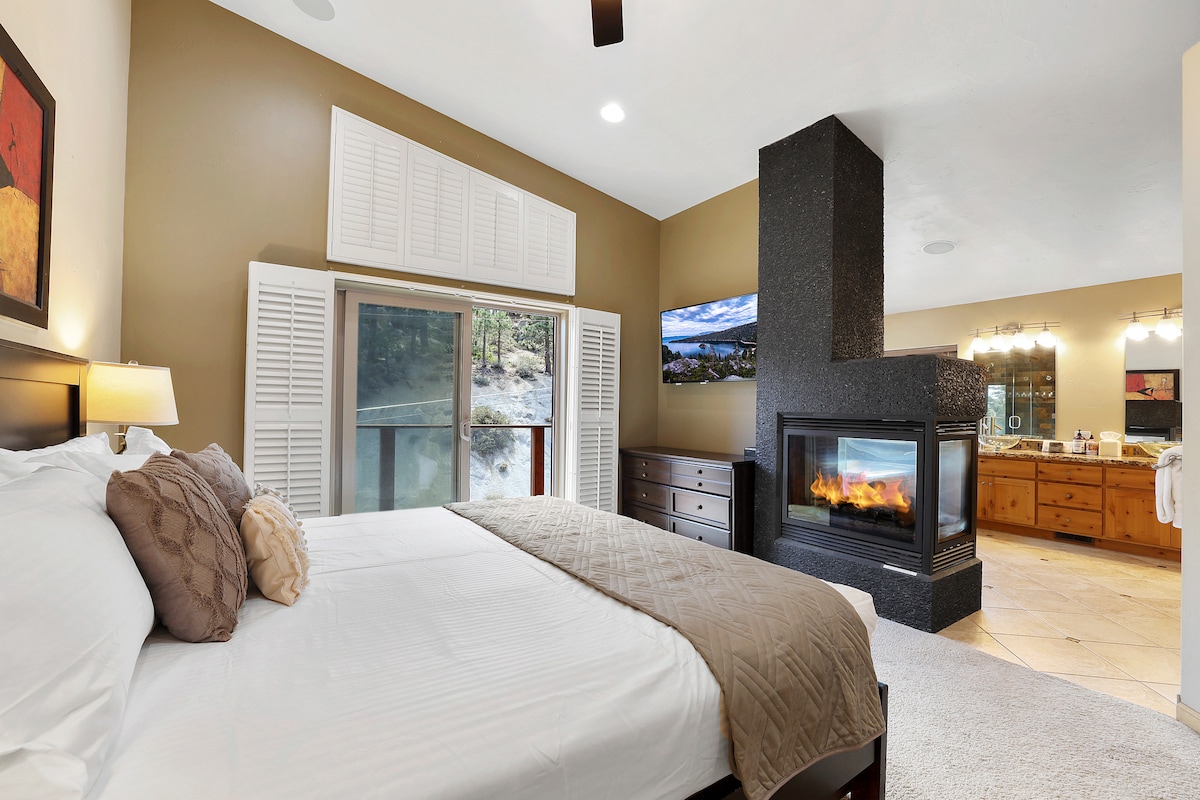 A spacious master bedroom features a large bed with luxury white bedding. A fireplace creates a cozy atmosphere beside the bed. Natural light fills the room through windows, with a view of the outdoors. An ensuite bathroom is visible, showcasing modern fixtures.