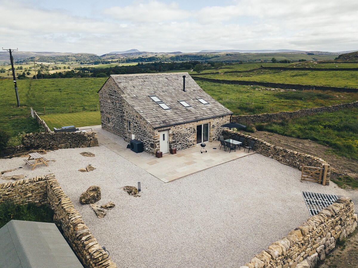 A stone barn conversion is set amidst a spacious gravel area, surrounded by natural landscapes. The building features a sloped roof with skylights, and a terrace with outdoor dining furniture. Rolling hills and scenic views form the backdrop, emphasizing the property's tranquil setting.