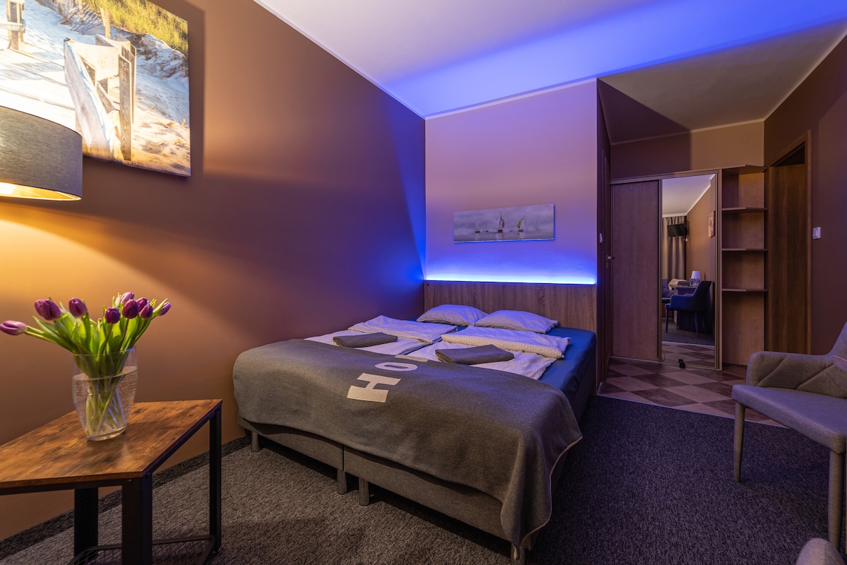 A cozy sleeping area features a spacious bed with light bedding arranged neatly. Soft purple lighting casts a warm hue on the walls, complemented by a framed nature scene above the bed. A small table holds a vase of fresh tulips, adding a touch of color.