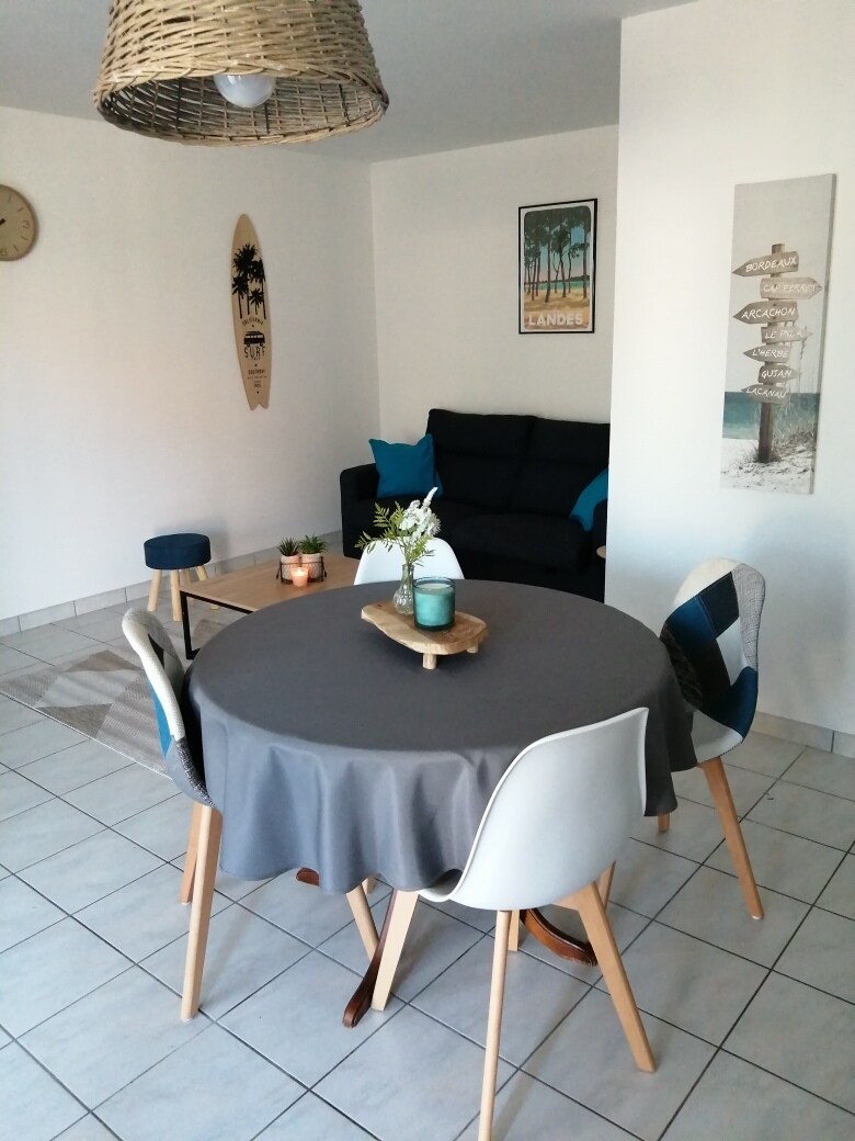 A dining area features a round table with a gray tablecloth, surrounded by four mixed-style chairs. In the background, a comfortable dark sofa is positioned against a light wall adorned with artwork, while a decorative surfboard adds a coastal touch to the space.