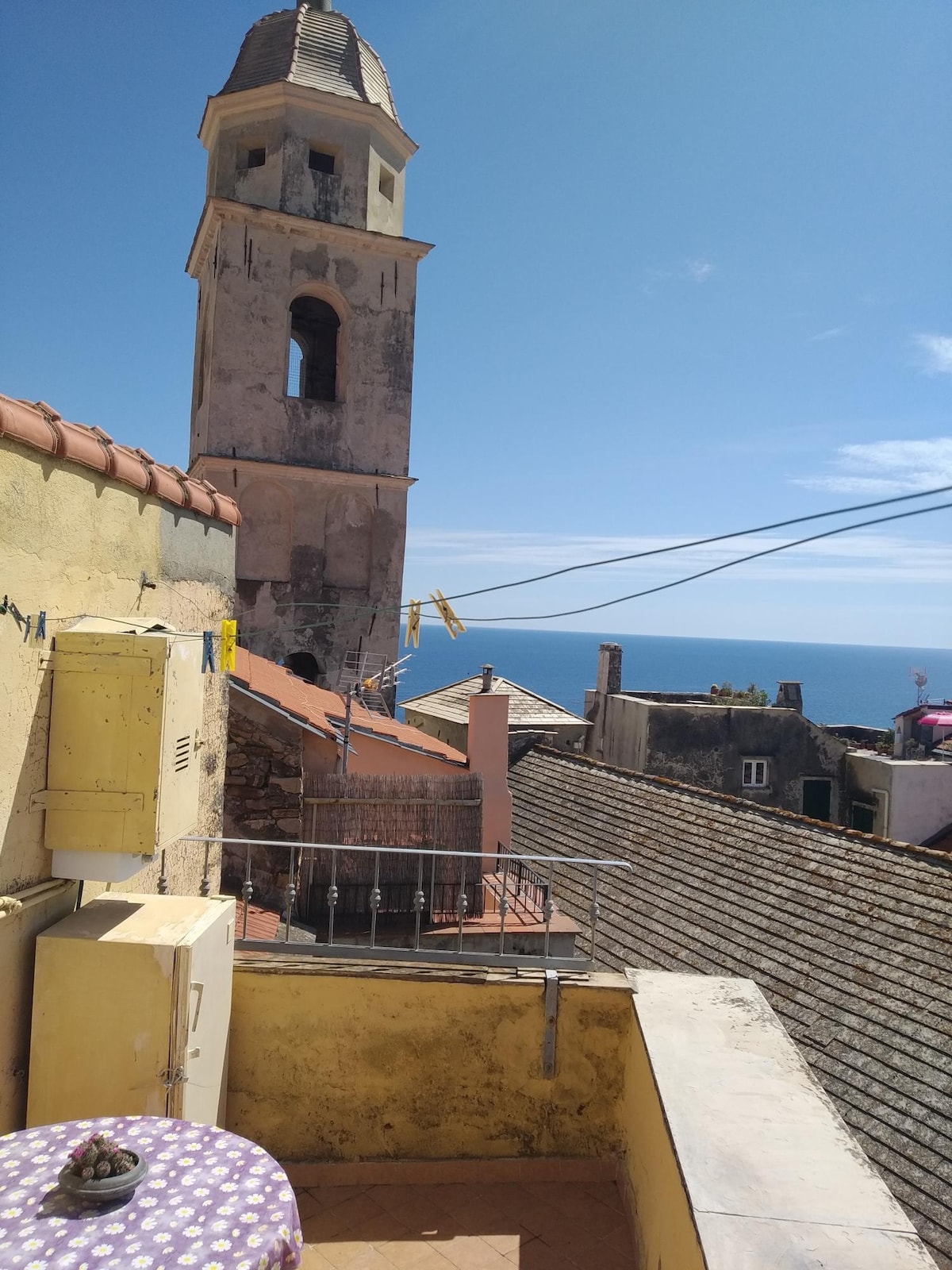A balcony reveals a view of the medieval village's rooftops, with a weathered bell tower in close proximity. The blue sea reflects the sky in the background, while laundry lines hang across the scene. A small table draped in a patterned cloth is positioned on the balcony.