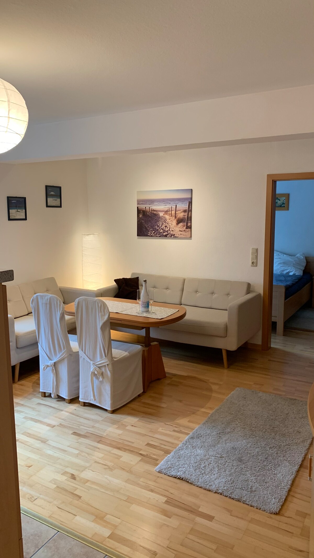 The image shows a welcoming living area with a light wooden floor and neutral-colored walls. A round dining table is surrounded by four upholstered chairs, while a comfortable sofa is positioned nearby. Soft lighting is provided by a standing lamp and wall art is displayed on one wall.