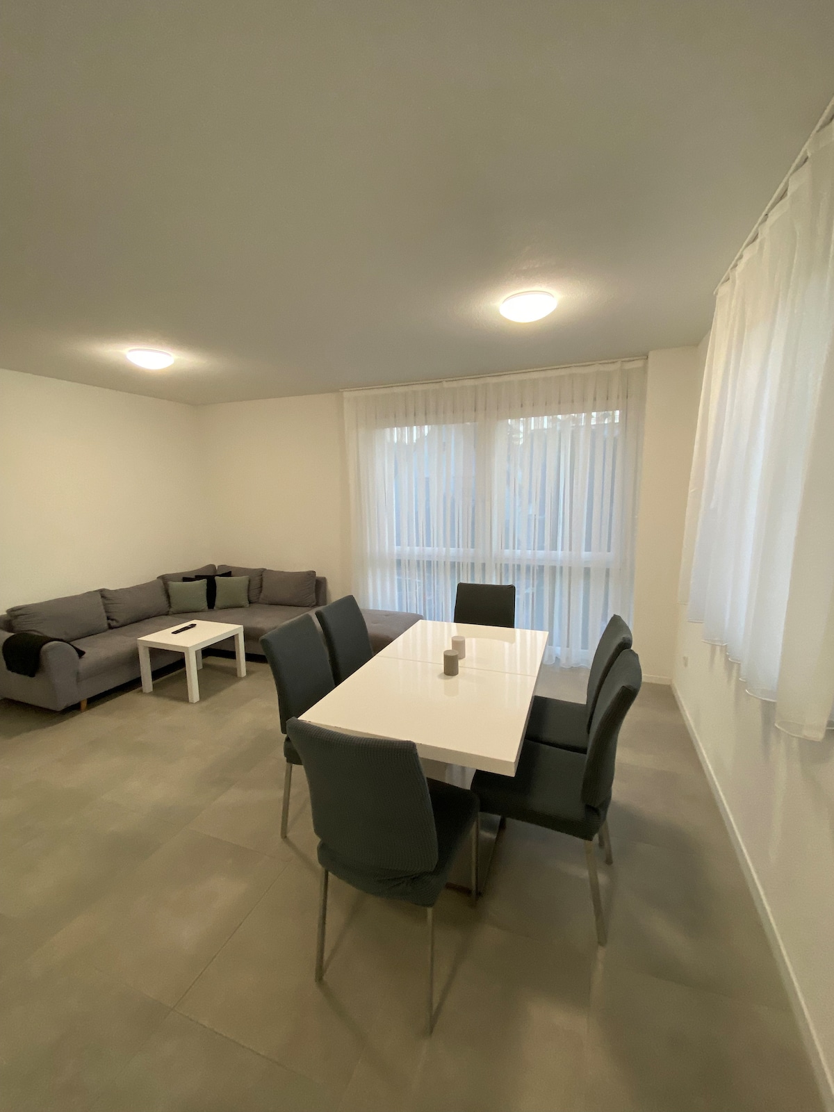 A modern dining area is visible, featuring a white table surrounded by six gray chairs. A comfortable gray sofa is positioned in a corner, complemented by natural light filtering through sheer curtains over a large window.
