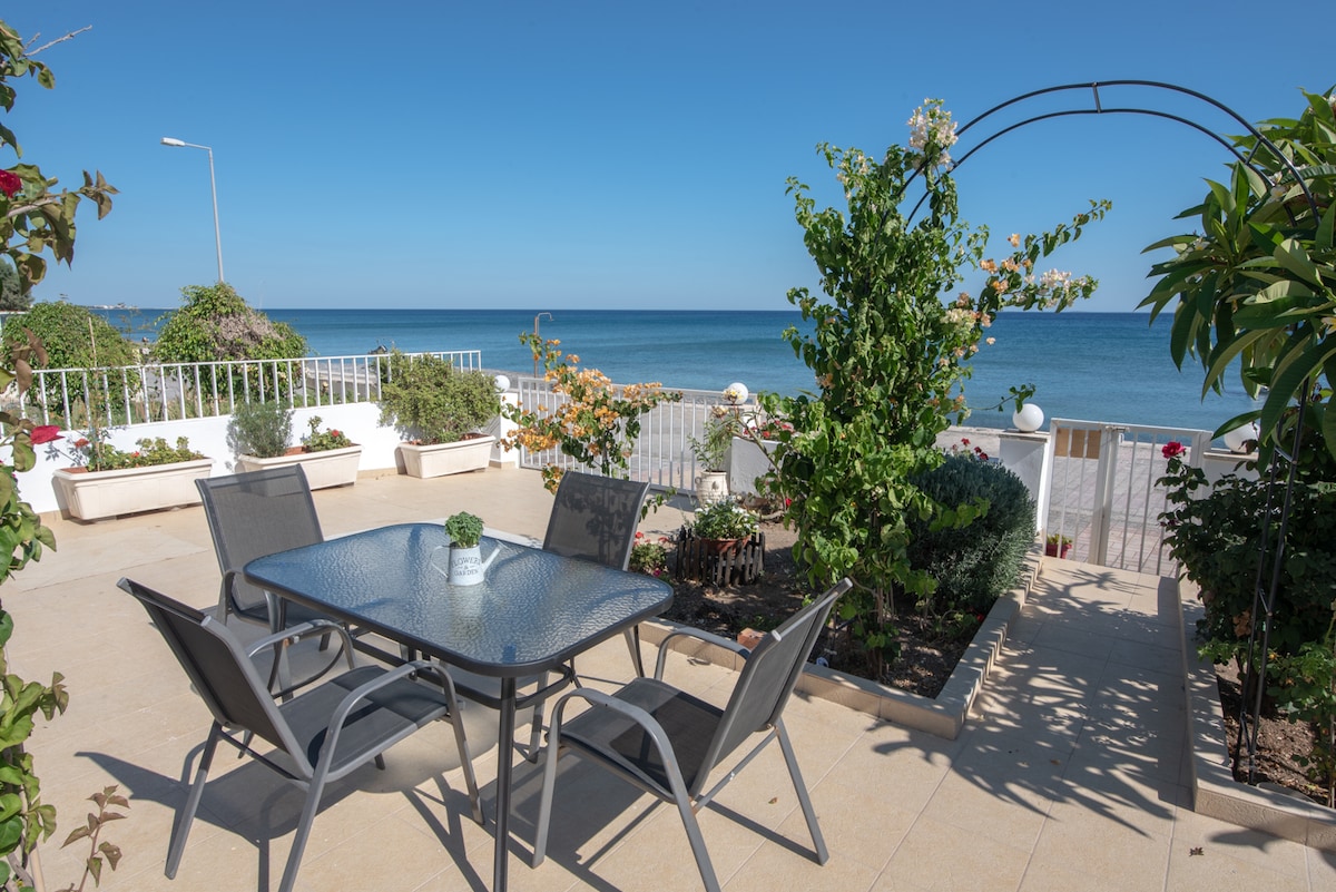 An outdoor terrace features a glass-top dining table surrounded by four chairs. Lush greenery and flowering plants enhance the space, while the serene sea is visible in the background. A trellis adds structure to the garden area, contributing to the inviting atmosphere.