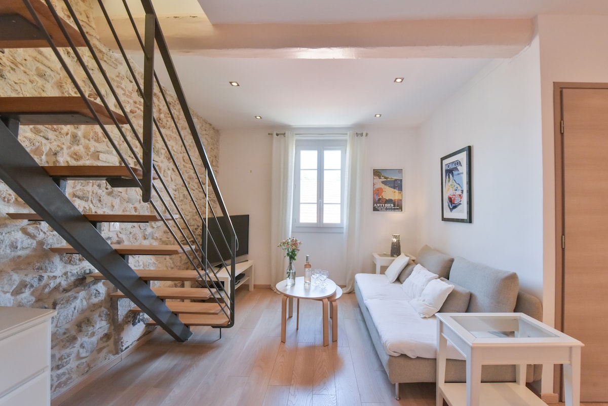 A spacious living area features a sleek staircase leading to the upper level. A comfortable sofa is situated adjacent to a round coffee table. Natural light fills the space from a window adorned with light curtains, while artwork adds character to the stone wall.
