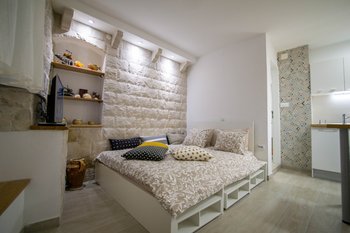 A spacious sleeping area features a low-profile bed adorned with various textured cushions in muted colors. A stone wall adds character to the room, while shelves display decorative items. Soft lighting accentuates the minimalist design and enhances the overall ambiance.