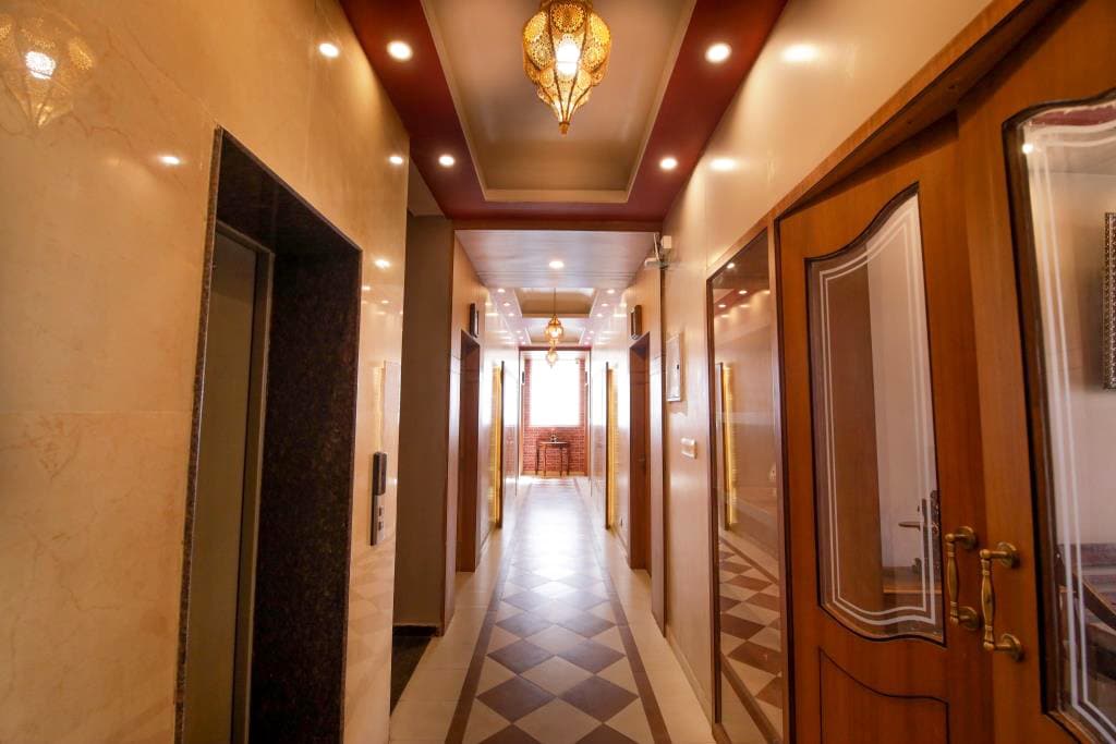 A well-lit corridor features stylistic lighting and decorative elements along the ceiling. Wooden doors line both sides, while a patterned floor leads to a welcoming area at the end. Elevators are positioned on one side, enhancing accessibility within the space.