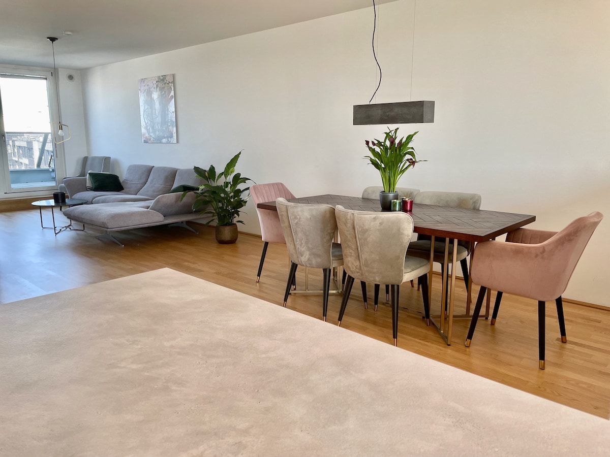 The spacious living area features a modern grey sofa and a large wooden dining table surrounded by soft, pastel-colored chairs. A potted plant adds a touch of greenery, while a minimalist light fixture hangs above the table, enhancing the room's inviting layout.