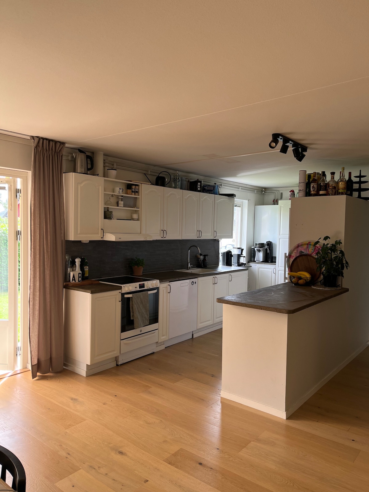 A modern kitchen is presented with white cabinetry and a sleek countertop. An island serves as a workspace, while essential appliances and utensils are organized neatly. Natural light enters through glass doors, illuminating the wooden floor and creating a functional and inviting cooking space.