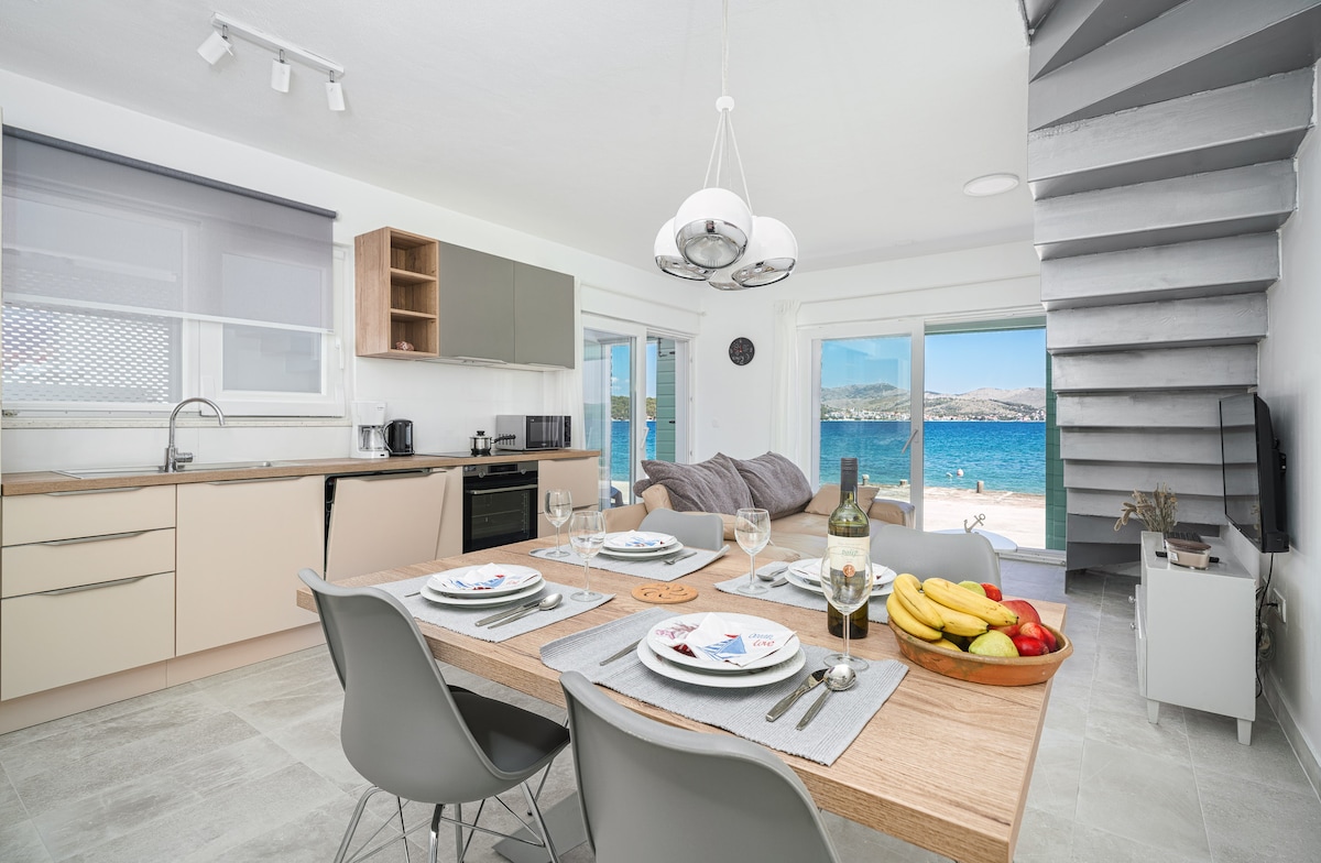 A modern kitchen and dining area are visible, featuring a wooden dining table set for four, with plates and glasses arranged neatly. The kitchen is equipped with sleek cabinets and appliances. Large glass doors lead to a view of the coastline, showcasing the sea and inviting natural light.