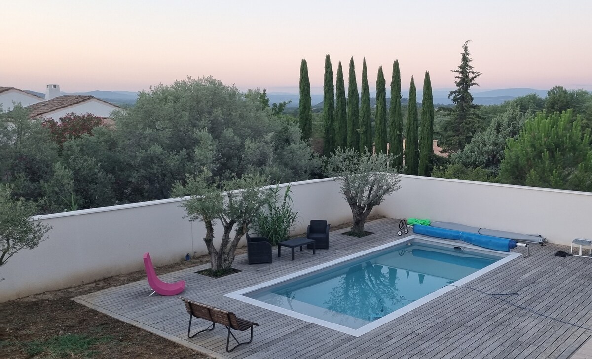 An outdoor area features a rectangular pool surrounded by wooden decking. Comfortable seating is arranged nearby, with olive trees and tall cypress trees providing natural greenery. A pink lounge chair and a blue sunshade are visible, complementing the serene landscape.