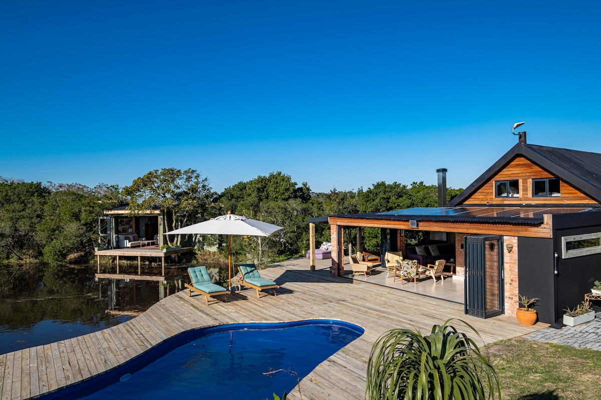 A serene outdoor space features a wooden deck surrounding a blue pool. The house boasts large windows and outdoor seating areas. An umbrella provides shade by the pool, while a small cottage sits nearby among lush greenery.