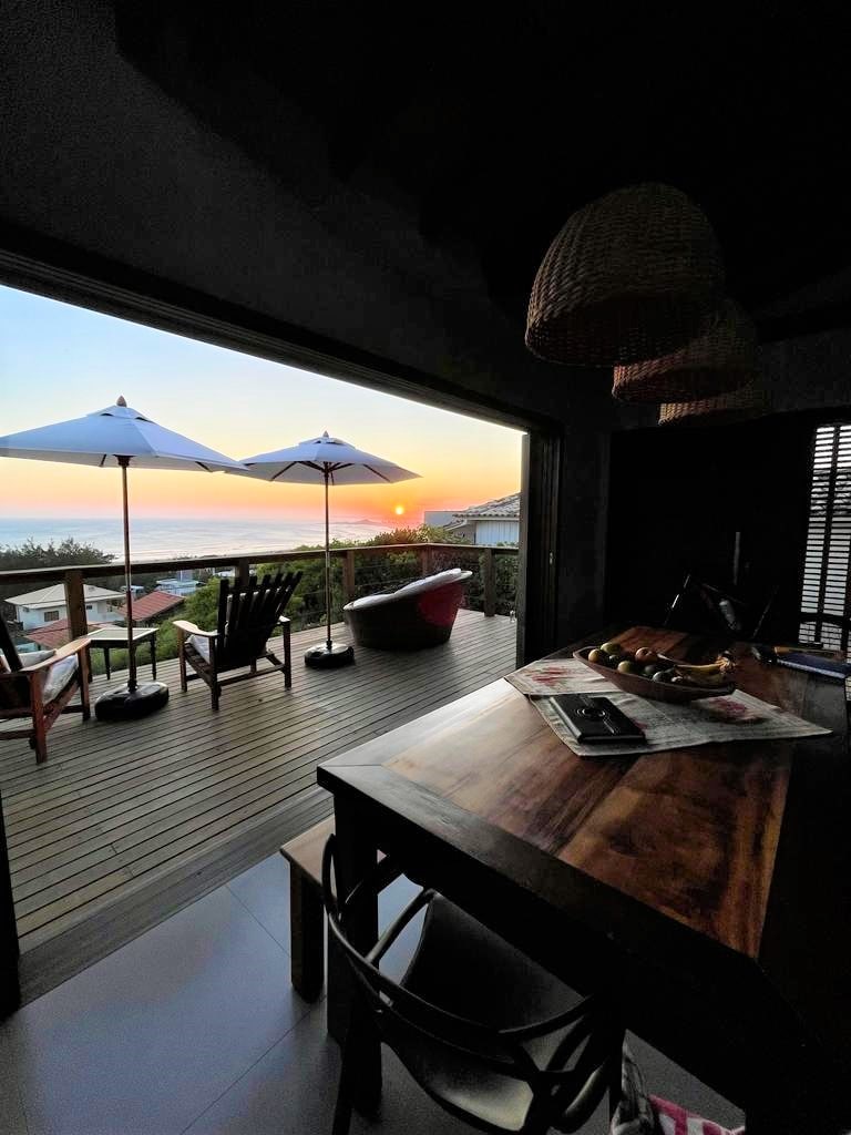 A cozy dining area features a wooden table and chairs, with sunlight streaming through large glass doors. Outdoor umbrellas shade lounge chairs on a deck offering a view of the sunset over the ocean. The scene is tranquil and inviting.
