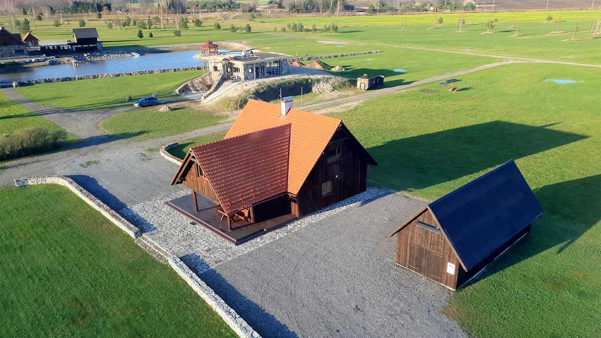 An aerial view of the villa showcases its wooden structure with a red tiled roof. Surrounding greenery and a gravel driveway are visible, along with additional buildings and a large body of water nearby, highlighting the spacious outdoor area.