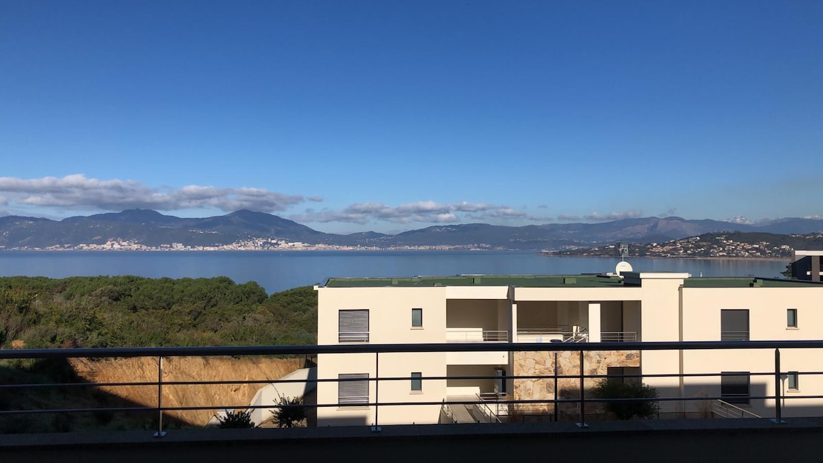 A panoramic view showcases the tranquil sea and distant mountains, with the coastline of Porticcio visible in the background. A modern building stands in the foreground, featuring clean lines and a minimalistic design.