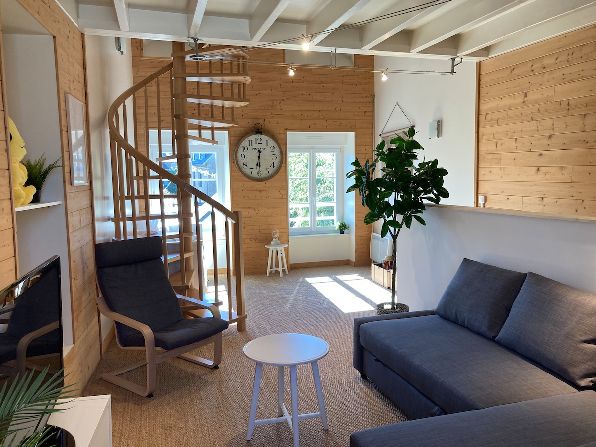 A well-lit living area features a spiral staircase leading to the upper levels. A sizable sofa and a minimalist chair provide seating. Natural light enters through multiple windows, and a large wall clock is prominently displayed, adding a functional element to the space.