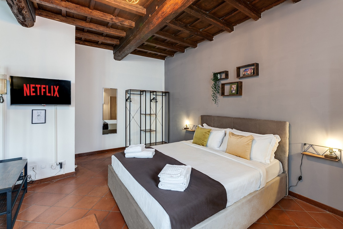 The spacious room features a comfortable bed with a neutral-colored coverlet and accent pillows. A wall-mounted SMART TV displays the Netflix logo. Exposed wooden beams accent the ceiling, while decorative frames enhance the light grey wall. A separate clothing rack and a full-length mirror are also present.