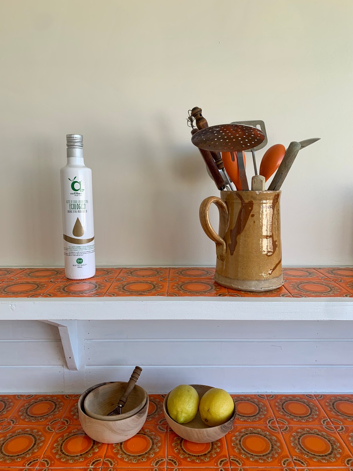 A wooden shelf displays a collection of kitchen essentials, including a ceramic jug and utensils. A bottle of oil is positioned beside a small wooden bowl containing a spoon, and another bowl holds two lemons, set against a patterned orange backdrop.