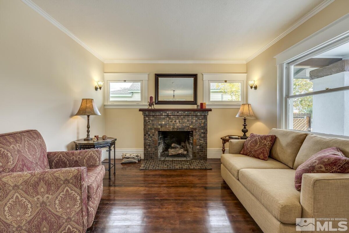 A living area is centered around a decorative fireplace, with a comfortable sofa and a patterned armchair placed on either side. Natural light is allowed in through the windows, highlighting the warm wood flooring and neutral wall colors.